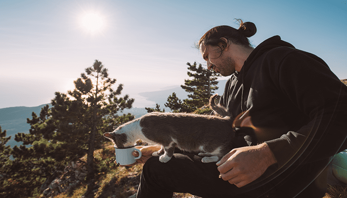 A middle-aged man gives his cat a drink of water from a white mug on the top of a mountain during their hike